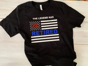 Black shirt. Wording at top states "the legend has retired.." Below is an american flag in white with a badge number in the starts in metallic red. In the middle of the flag is the word "retired" in bold blue vinyl